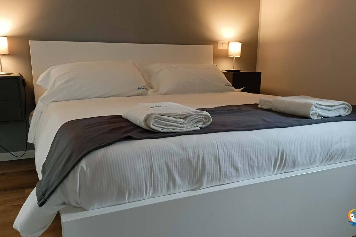 Ein ordentlich bezogenes Bett mit weißen Leinentüchern, ein gefaltetes Handtuch und eine dunkle Decke. Zwei Nachttische mit Lampen, und ein Logo an der Wand.