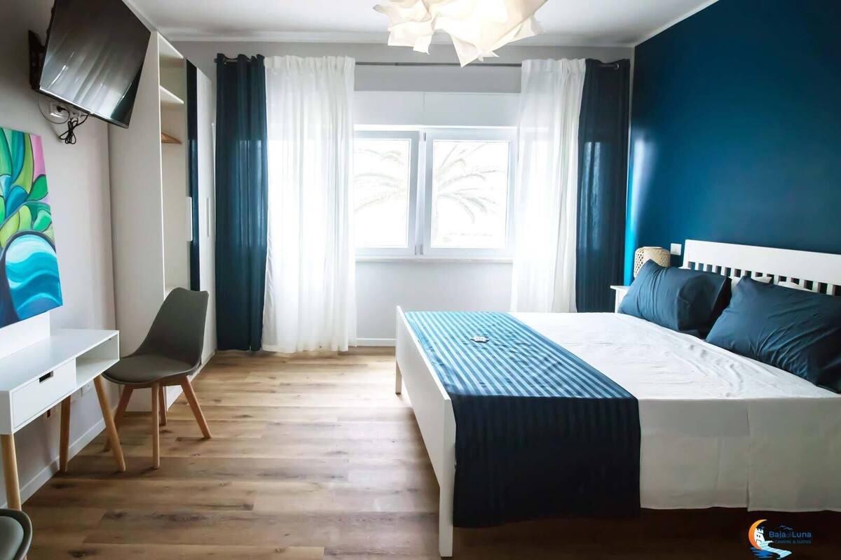 Ein Schlafzimmer mit Bett, Schreibtisch, Stuhl, Fernseher und einem Fenster mit Vorhängen.