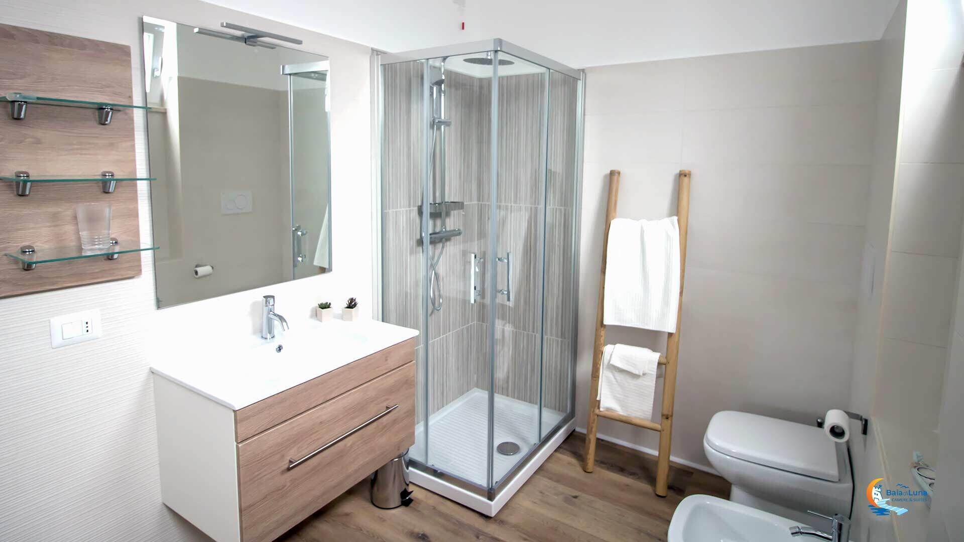 Un baño moderno con ducha de vidrio, un lavabo blanco con tocador de madera, un espejo grande y un inodoro con toallero.