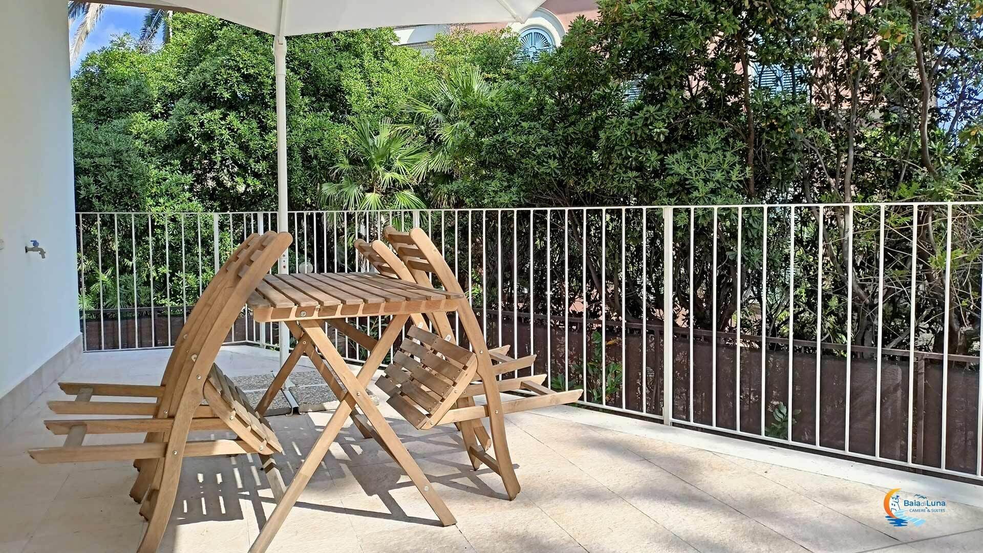 A wooden outdoor seating area with a white metal railing, surrounded by lush greenery.
