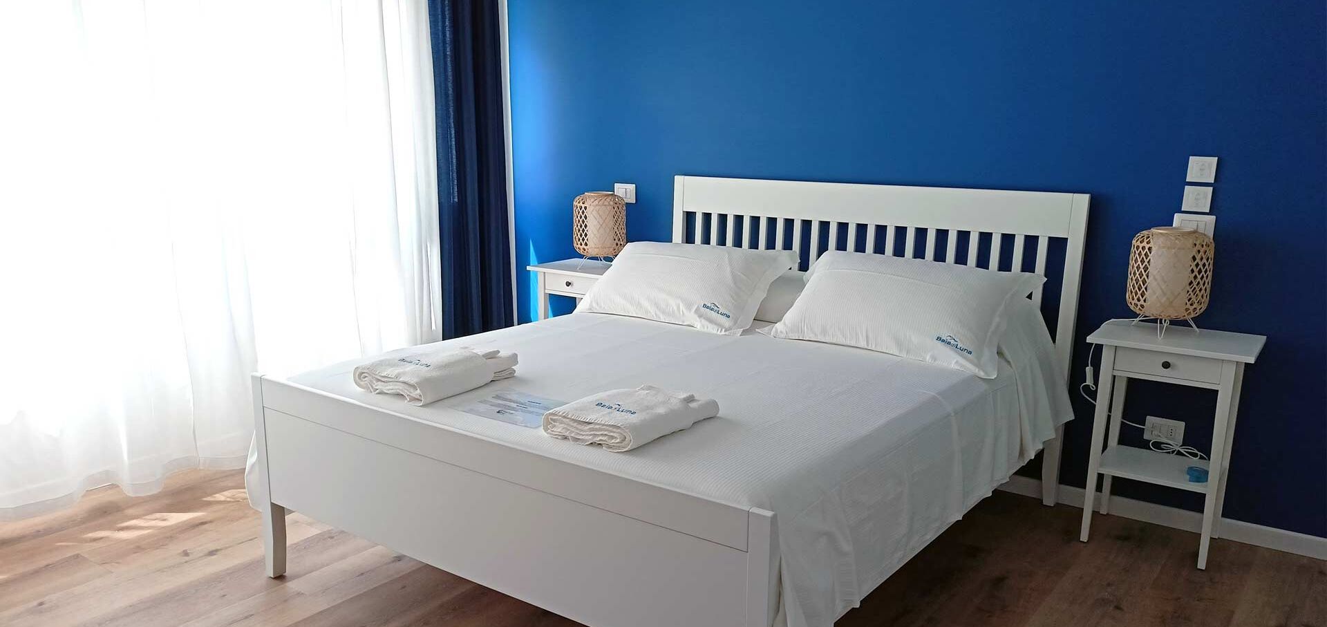 Una cama bien tendida con sábanas y almohadones blancos, dos mesitas de noche con lámparas y una ventana con cortinas traslúcidas.