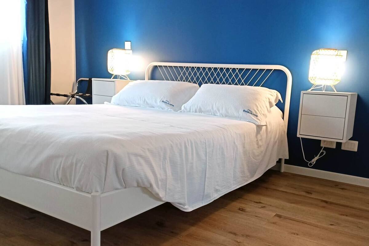Ein Bett mit weißer Bettwäsche, einem weißen Metall-Kopfstück und zwei Nachttischen mit Lampen.