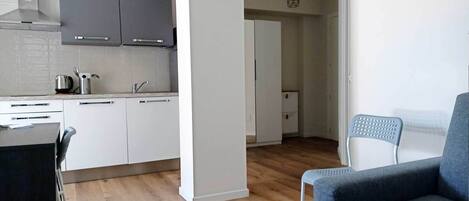 A modern kitchen with white cabinets, a stainless steel range hood, and a wooden floor. There is a dining area with a blue chair and a white table.