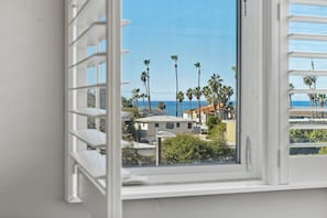 A window with white blinds, a view of a coastal area with palm trees and a building with a red roof.
