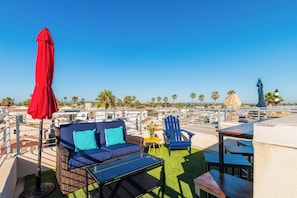Een dakterras met buitenmeubilair, waaronder een bank, stoelen en tafels, met uitzicht op een kustgebied met palmbomen en gebouwen.