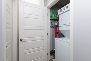 A laundry room with a white door, a washing machine, a dryer, and cleaning supplies on shelves.