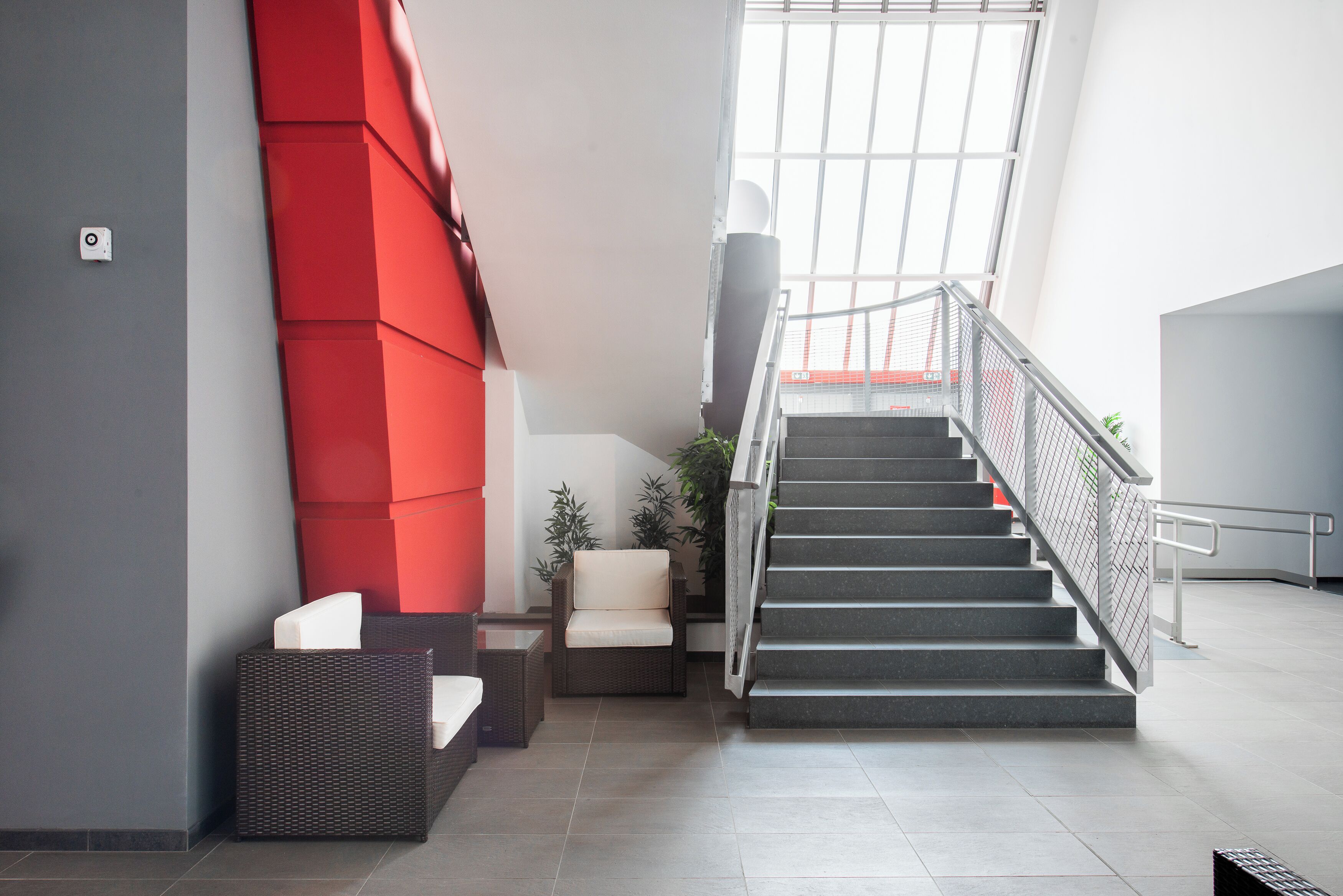 A modern interior with a staircase, a red accent wall, and a seating area with a wicker sofa and a chair.