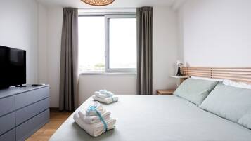 Ein ordentlich bezogenes Bett mit Handtüchern, ein Fernseher auf einer Kommode und ein Fenster mit Vorhängen.