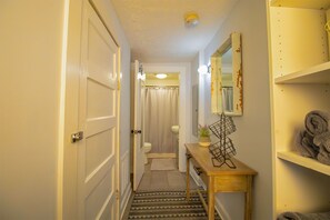 Un couloir étroit mène à une salle de bain équipée de toilettes, d’un rideau de douche et d’une petite table sur laquelle se trouvent une plante et un porte-serviettes.