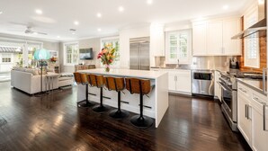 A modern kitchen with a central island, bar stools, stainless steel appliances, and a dining area with a sofa and TV.