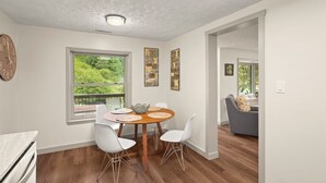 A modern dining area with a round table, white chairs, and a view of greenery through a large window. - Cozy riverfront cabin on the New River.  Close to Boone and Appalachian State. (Boone)