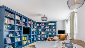 A modern living room with a large blue bookshelf, a round dining table with a glass top, and a herringbone wood floor. - Stylish Flat Close To Beach - Pass the Keys (Chichester)