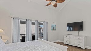 Une chambre avec un grand lit, un ventilateur de plafond, une télévision, une commode blanche et un balcon avec des rideaux.