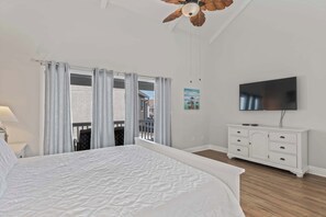 Une chambre avec un grand lit, un ventilateur de plafond, une télévision, une commode blanche et un balcon avec des rideaux.