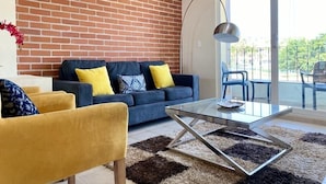 A modern living room with a brick wall, a blue sofa with yellow cushions, a glass coffee table, and a view of the outdoors through large windows. - SM Apartments (San José del Cabo)