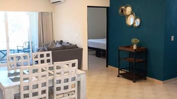 A room with a dining table, chairs, a sofa, a bed, a wall-mounted mirror, and a small shelf with a vase.
