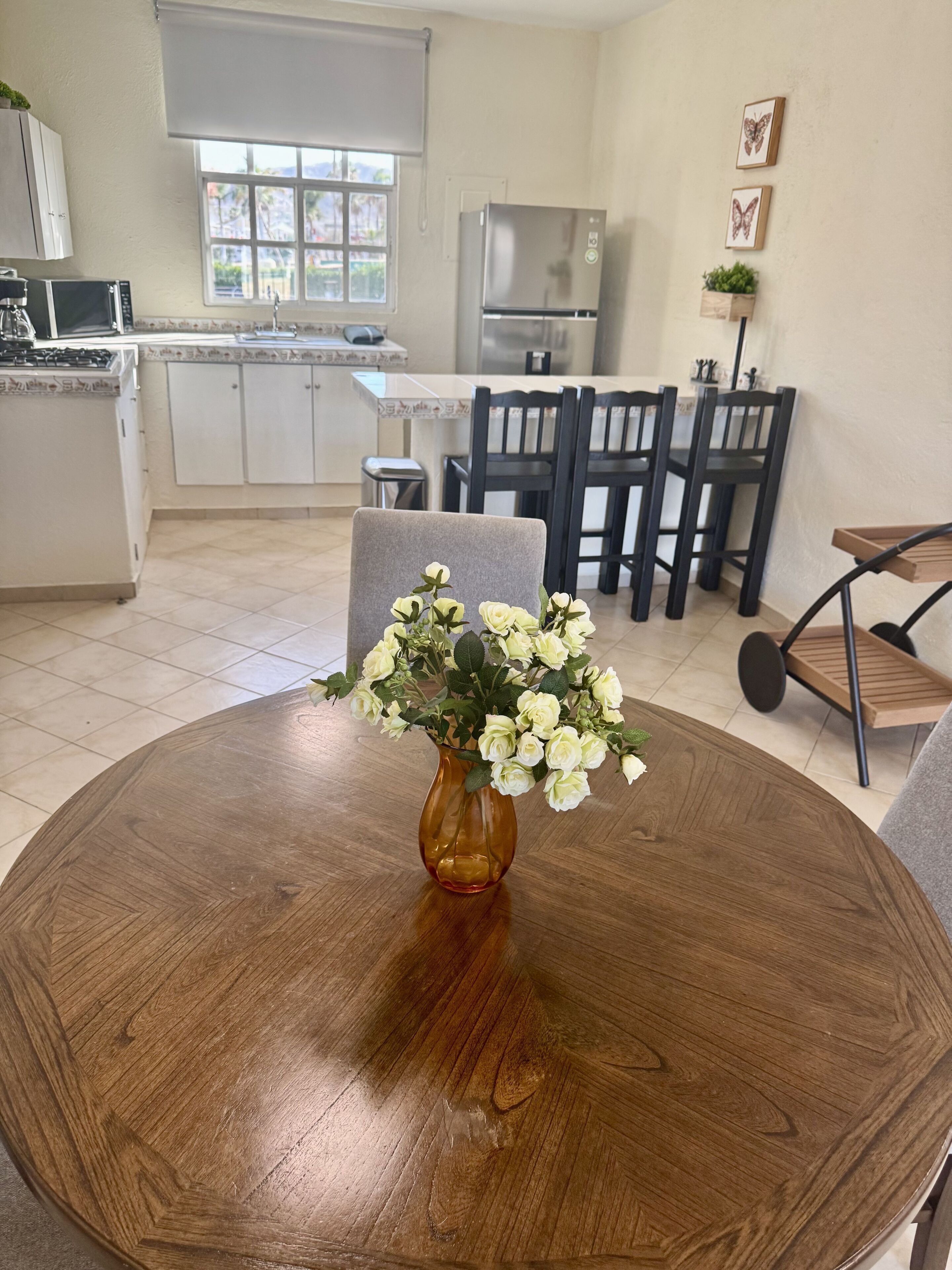 Een eetgedeelte met een ronde houten tafel, stoelen, een vaas met bloemen, een keuken op de achtergrond en een raam met uitzicht.