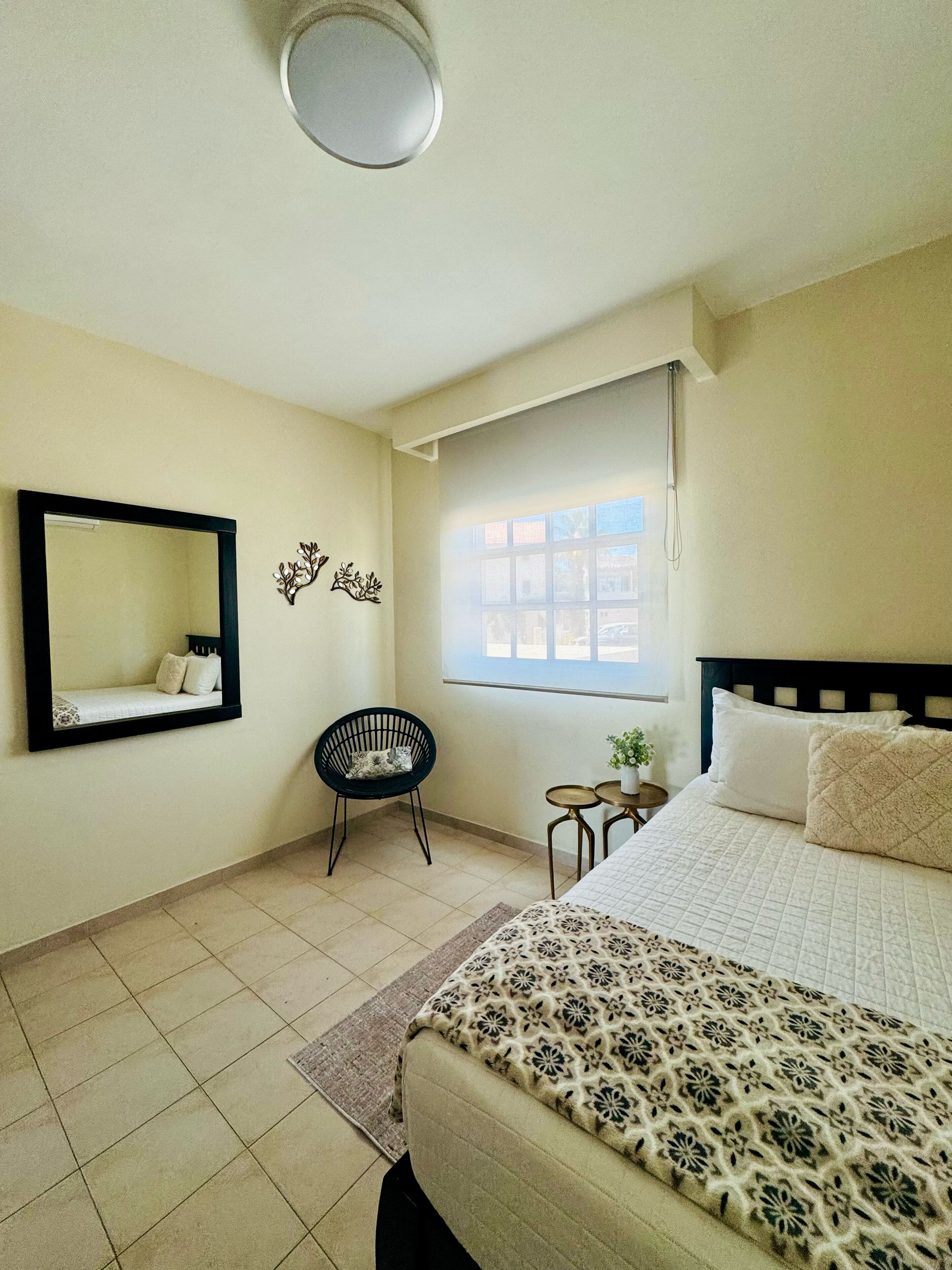 A bedroom with a bed, a chair, a small table, a window, and a mirror on the wall.