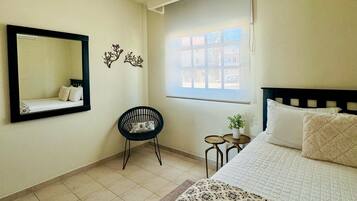 A bedroom with a bed, a chair, a small table, a window, and a mirror on the wall.