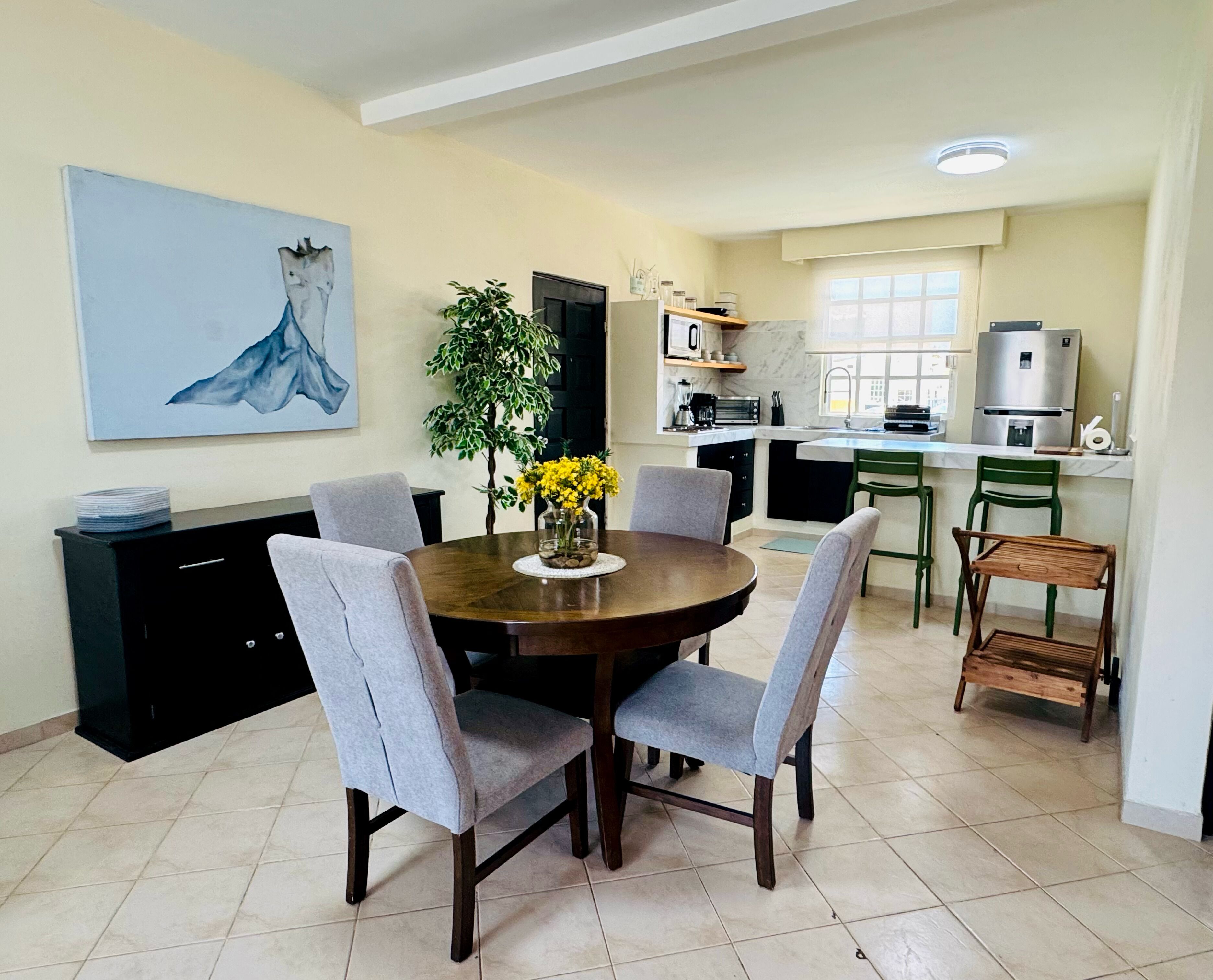 A dining area with a round table, grey upholstered chairs, and a vase of yellow flowers. There is a kitchen in the background with modern appliances and a window.