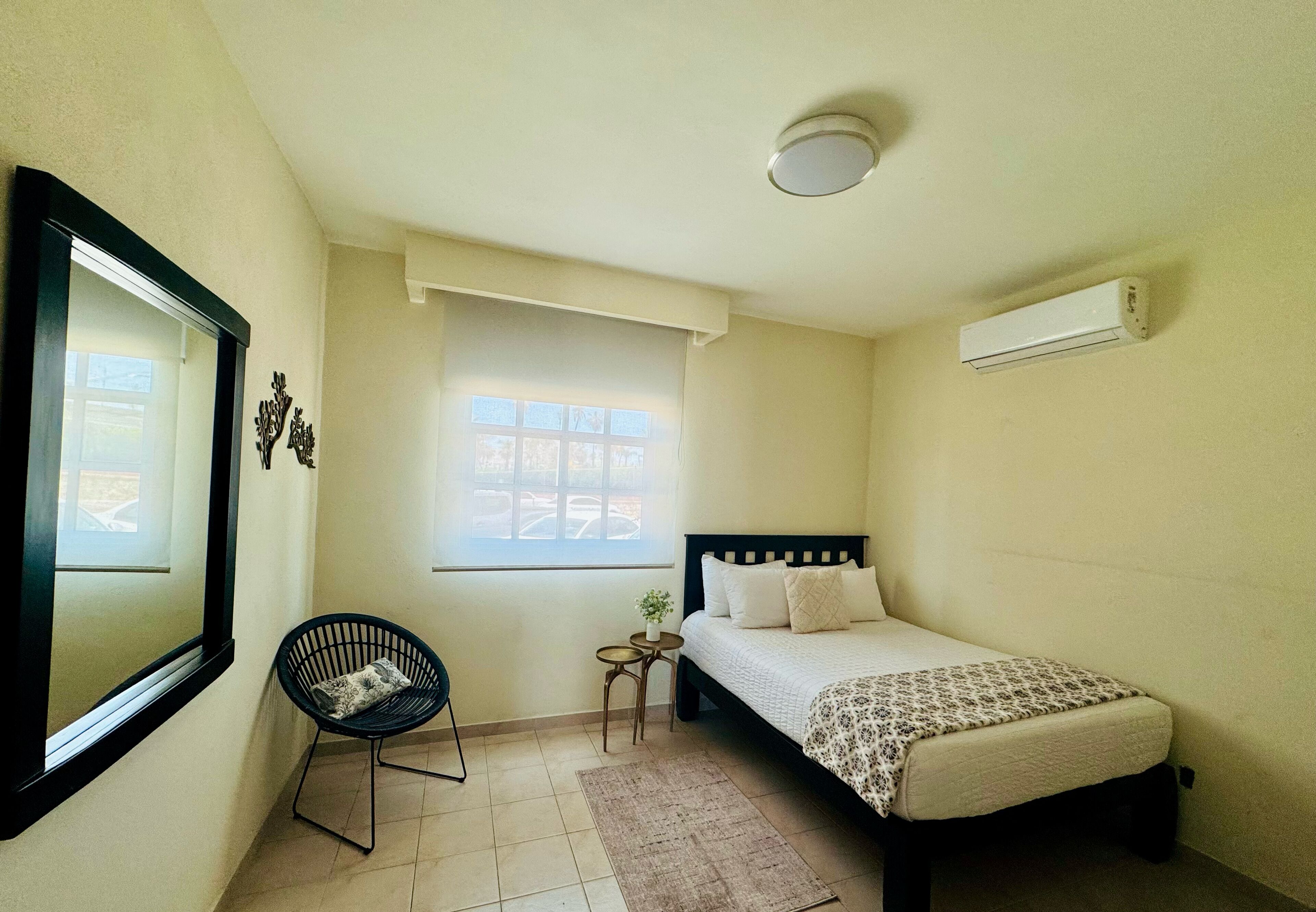 Un dormitorio con cama, una silla, una mesita, un ventanal y un equipo de aire acondicionado.