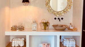 A bathroom vanity with a gold-framed mirror, a hanging light, and shelves with toiletries and a magazine. - SM Apartments (San José del Cabo)