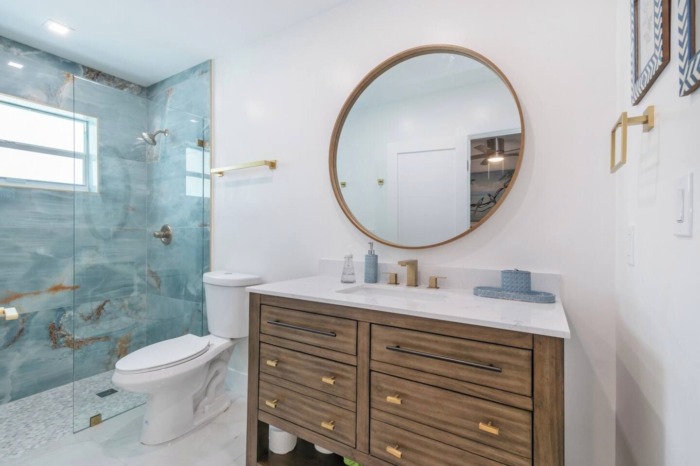 A bathroom with a glass shower enclosure, a white toilet, a wooden vanity with drawers, a round mirror, and a ceiling fan.
