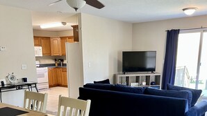 Ein Wohnzimmer mit einem dunkelblauen Sofa, einem Holzesstisch mit Stühlen, einem Deckenventilator und einem Fernseher auf einem Schrank.