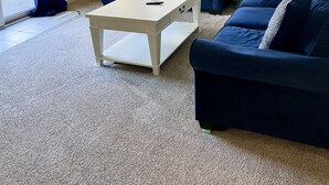 Ein Wohnzimmer mit beigefarbenem Teppich, zwei blauen Sofas, einem weißen Couchtisch und einem Fenster mit Jalousien.