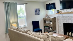 Ein Wohnzimmer mit einem beigen Sofa, einem blauen Sessel, einem Flachbildfernseher, einem Kamin und einem Fenster mit Vorhängen.