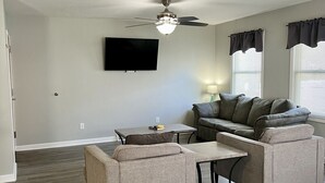 A living room with a ceiling fan, a flat-screen TV, a sofa, a coffee table, and a window with curtains. - Seascape Townhome #69- Sunset Vacation Rentals (Geneva)