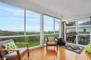 A spacious balcony with large windows offering a view of the sea and greenery. - Olde Beach Pearl - Waikanae Beach Holiday Home (Waikanae)