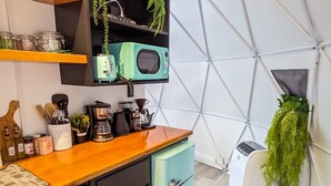 A kitchen with a wooden countertop, a teal refrigerator, and a microwave on a shelf. There are utensils in a container, a coffee maker, and a plant hanging on the wall.