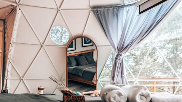A cozy bedroom with a geometric-patterned wall, a bed with dark bedding, a round mirror, and a window with curtains.