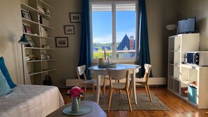 A room with a bed, a dining table with chairs, a bookshelf, and a window with a view of buildings. - Le Saint Merry apartment in Fontainebleau in a quiet, central location. (Fontainebleau)