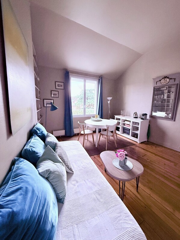 A modern living room with a white sofa, a dining table with chairs, a coffee table with flowers, and a bookshelf.