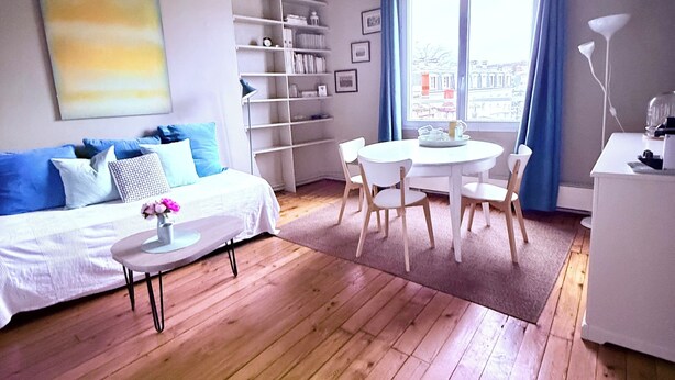 A modern living room with a sofa, a dining table with chairs, a bookshelf, and a large window with curtains. - Le Saint Merry apartment in Fontainebleau in a quiet, central location. (Fontainebleau)