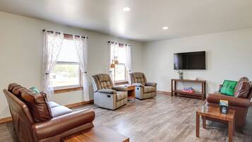 A spacious living room with leather sofas, a wooden coffee table, and two recliner chairs. A flat-screen TV is mounted on the wall, and a small table with a plant and a box is visible.