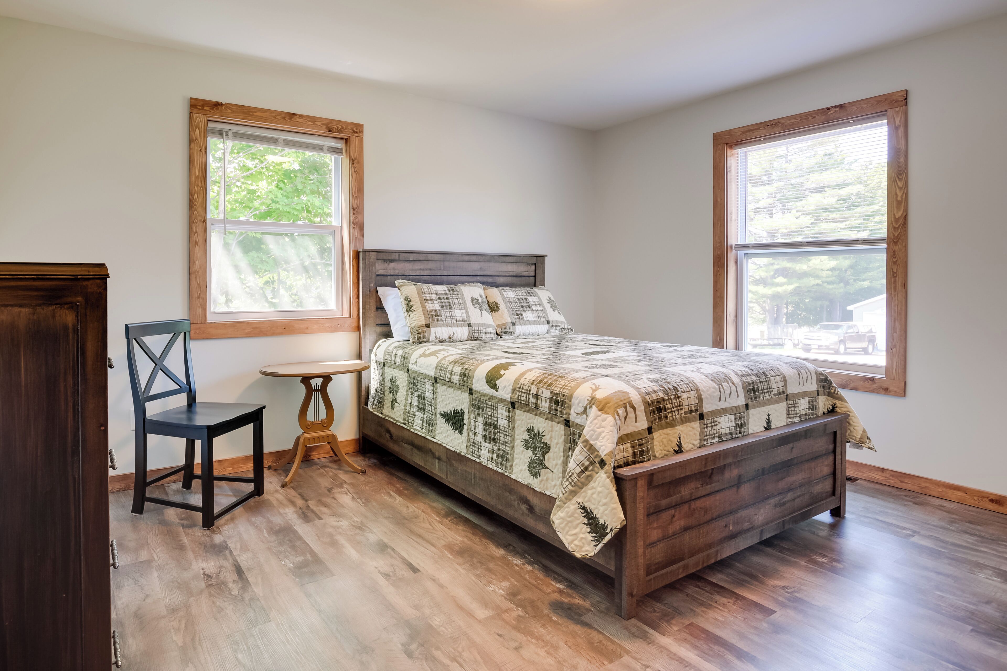 A bedroom with a wooden bed, two windows, a chair, and a small table.