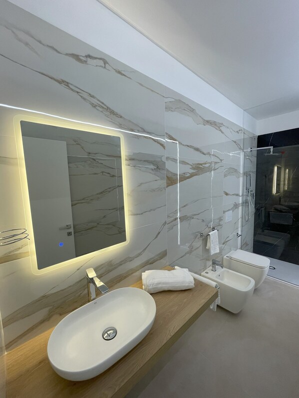 A modern bathroom with a marble backsplash, a large mirror, a sink, a bidet, and a glass shower enclosure.