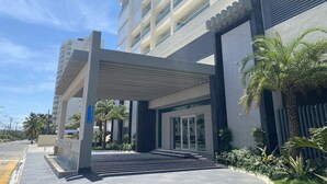 Entrada de hotel moderna com galeria coberta, palmeiras e céu claro.