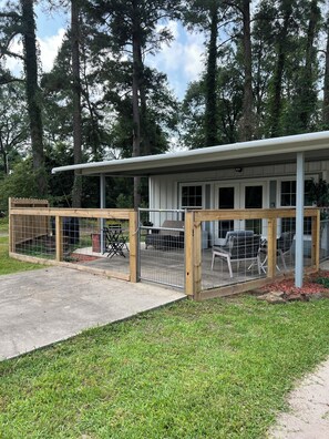 A cabin with a covered patio, a fenced-in area with outdoor furniture, and a view of tall trees in the background.
