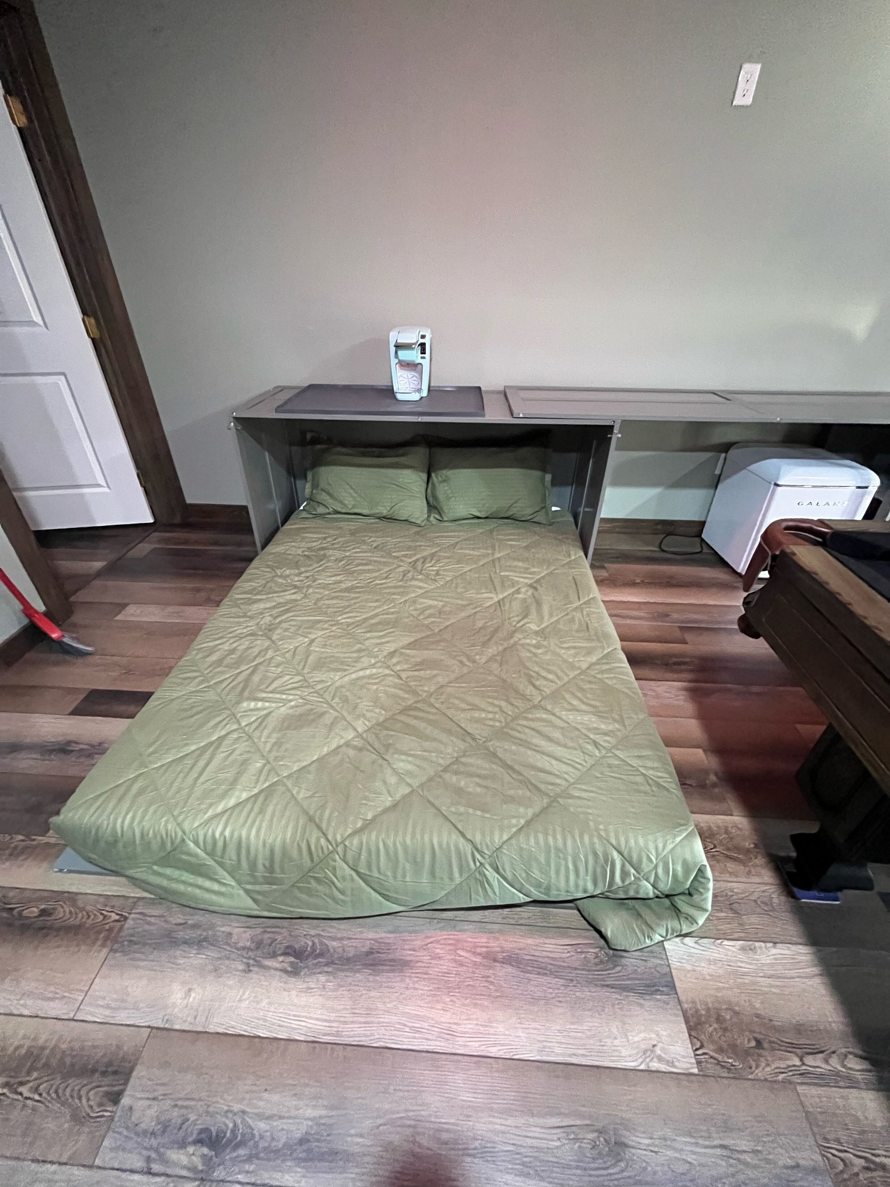 Uma cama de casal com um edredom verde, uma mesinha de cabeceira com uma luminária e uma pequena mesa com um aparelho branco.