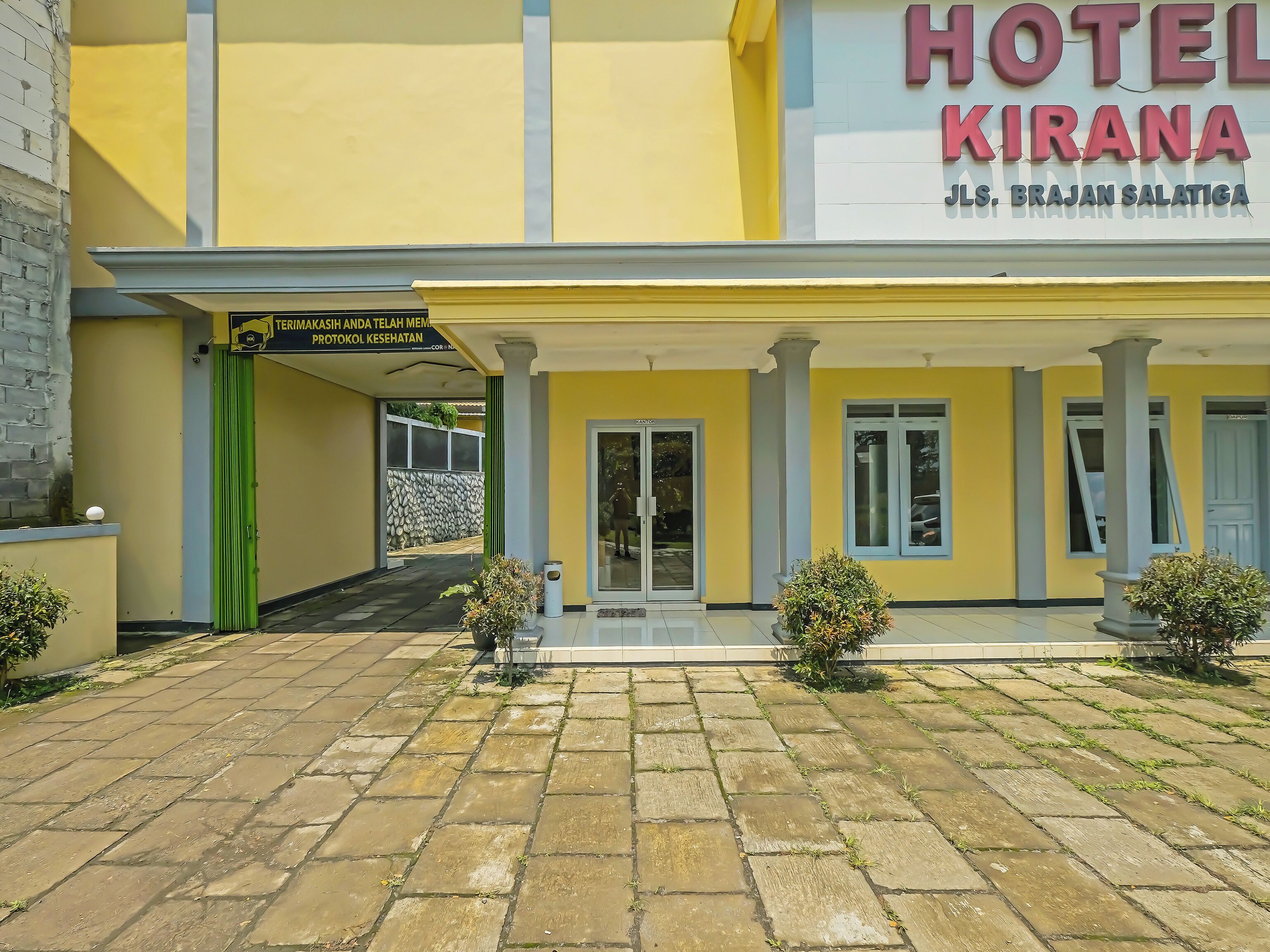 Fachada amarilla de un hotel, con un cartel que dice "HOTEL KIRANA" y un acceso cubierto que lleva a la entrada.