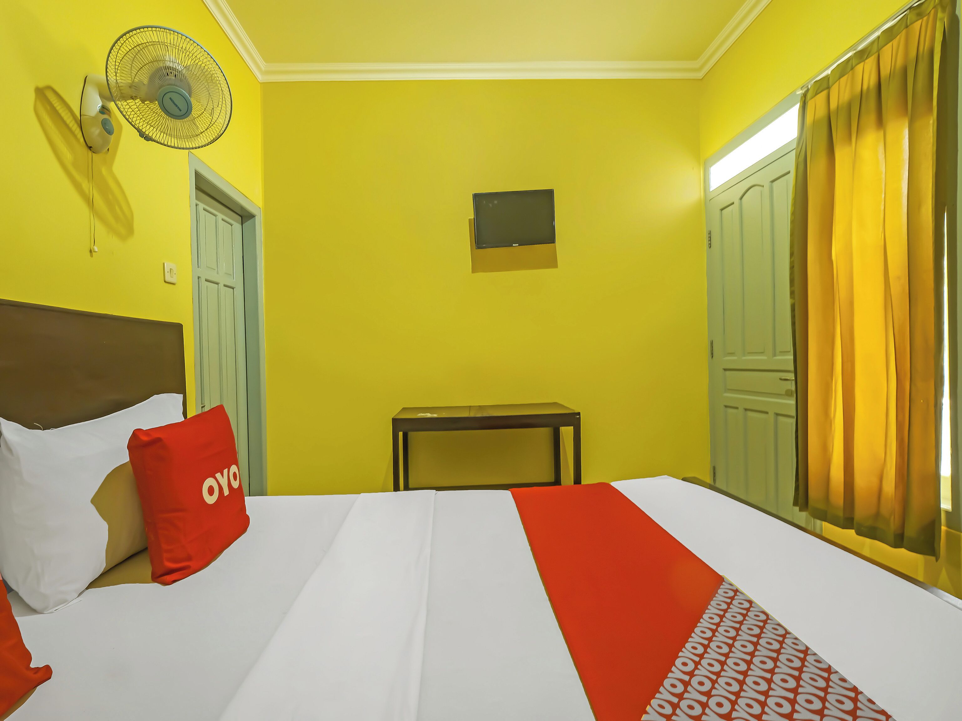A hotel room with a bed, a small table, a wall-mounted fan, and a door.