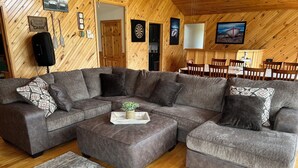 Interior acogedor de una cabaña con un amplio sofá modular, techo de madera y comedor con mesa y sillas.