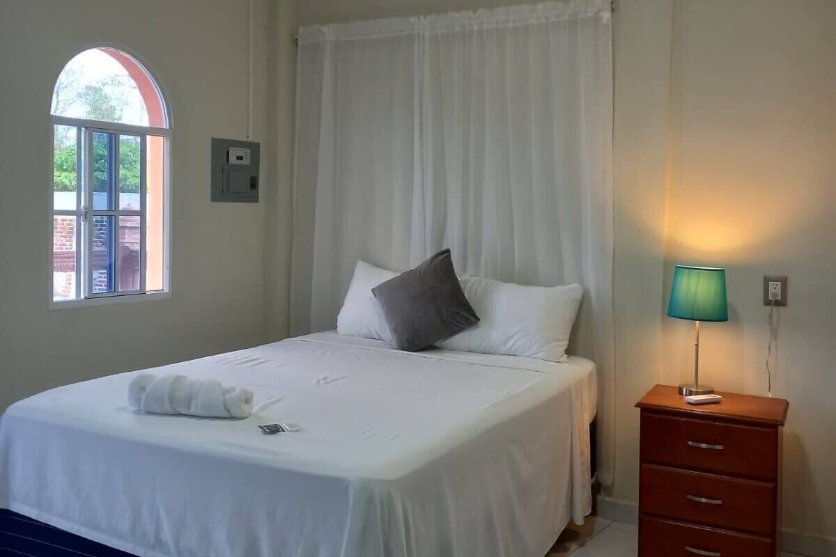 A bedroom with a bed, a nightstand with a lamp, a window with curtains, and a view of the outside.
