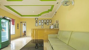 Un hall d’hôtel avec un plafond vert et blanc, un réfrigérateur vert, une table basse en verre, un canapé vert clair et un mur orné de cadres contenant des certificats.