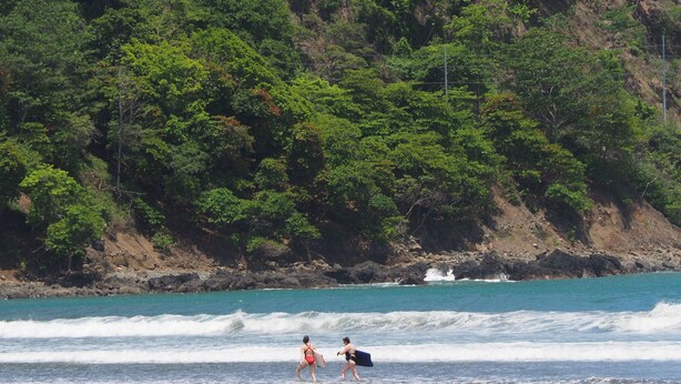 A beach with waves, a rocky shoreline, and dense green foliage in the background. - Family Paradise Macaws 3 (Jacó)