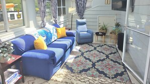 Una habitación iluminada por el sol con un sofá azul, una silla, un tapete estampado y plantas en macetas.
